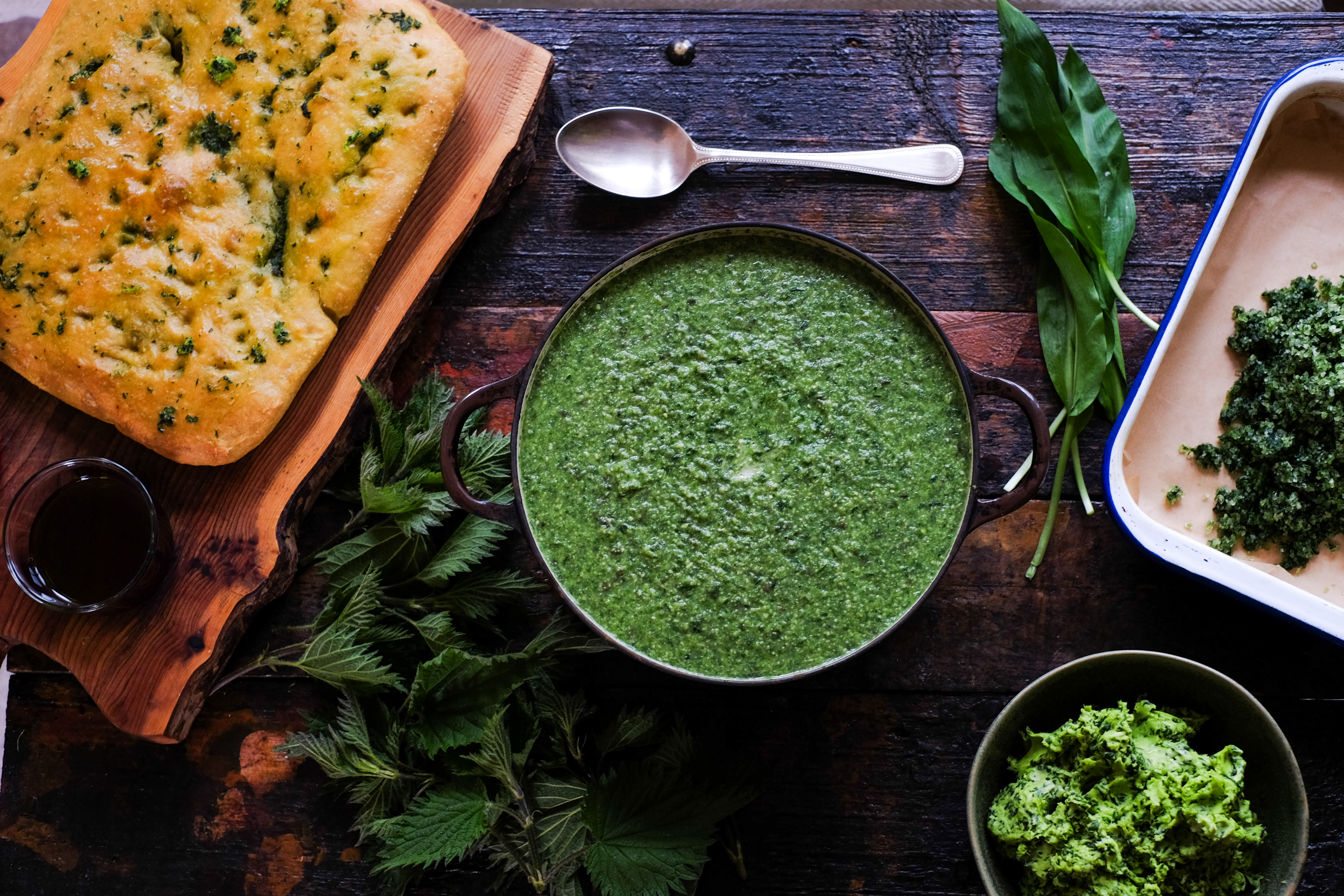 A wooden surface displaying a bowl of green soup, a piece of herb bread, fresh greens, and a small dish of chopped herbs.