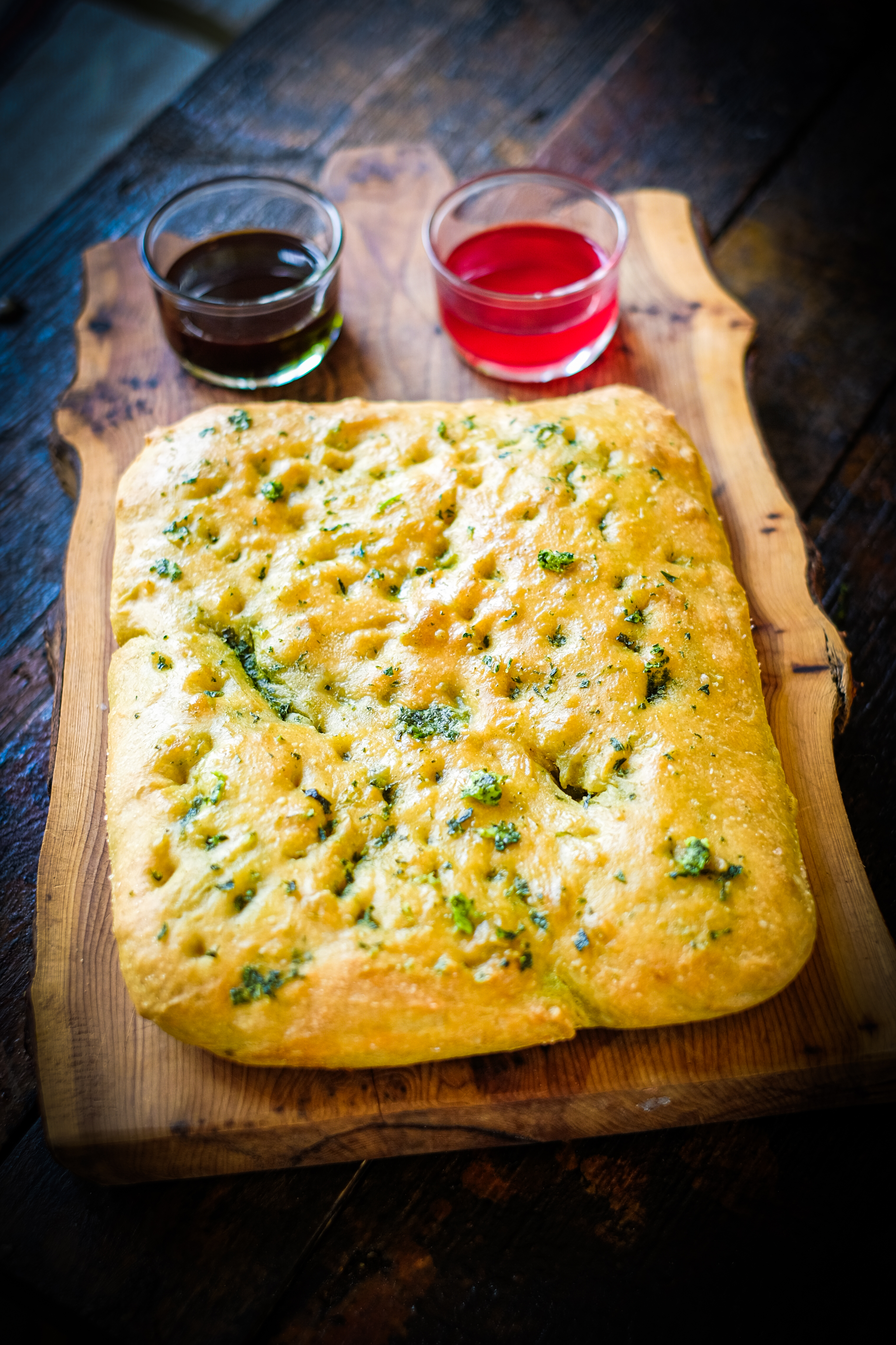A freshly baked flatbread with herbs, served on a wooden board, accompanied by two small bowls of sauces, one dark and one red.