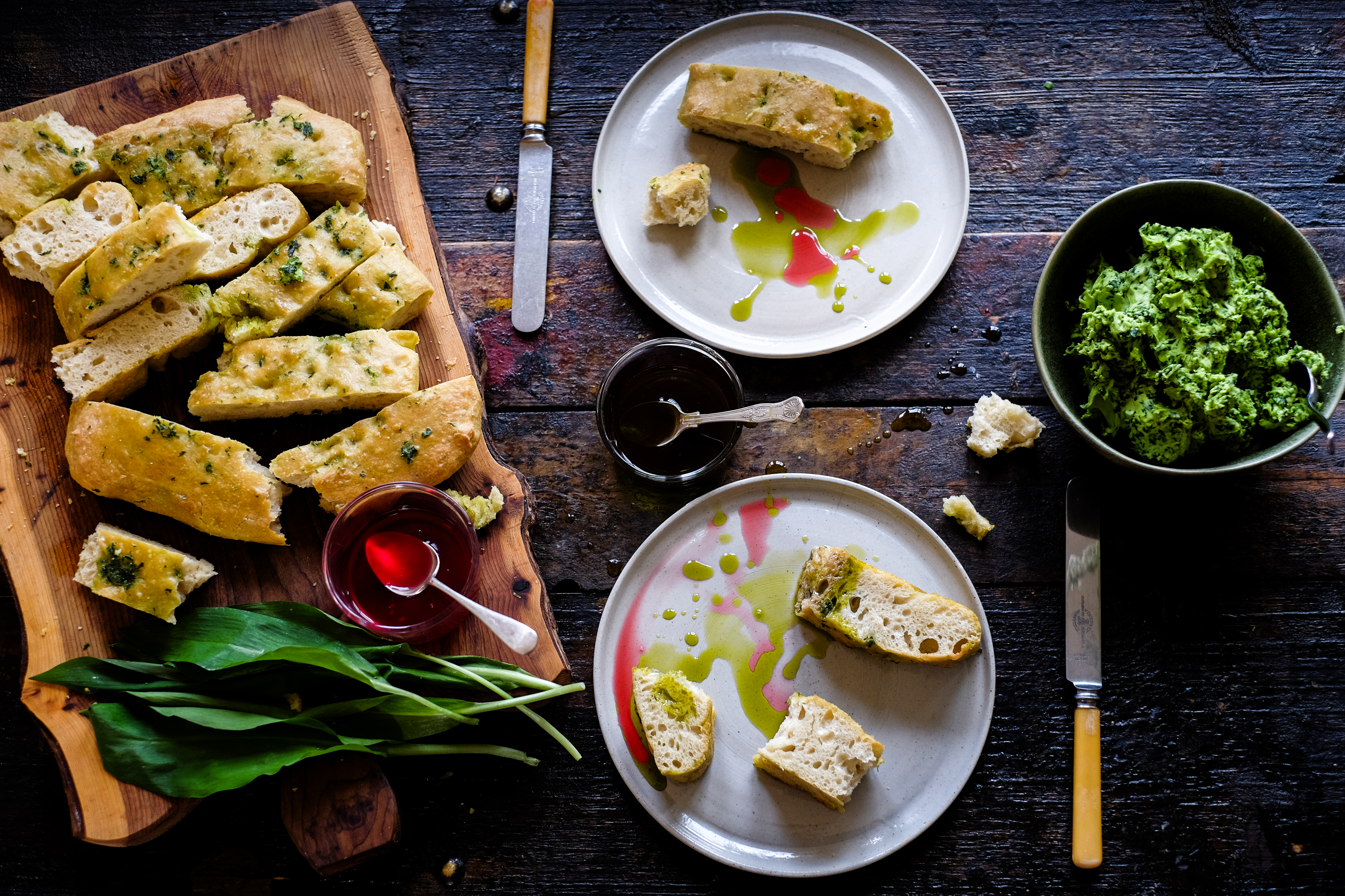 Freshly sliced herb bread on a wooden board with green sauce, accompanied by two plates of bread pieces drizzled with oil and a bowl of green spread.